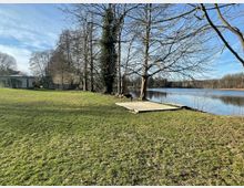 Grüne Wiese mit mehreren laublosen Bäumen am Ufer eines ruhigen Sees; im Vordergrund eine kleine Holzplattform und im Hintergrund ein Gebäude mit weißer Fassade sowie ein Zaun. Der Himmel ist klar und blau.