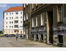 Straßenszene mit einem grauen, verwohnten Gebäude rechts, dessen Fassade mit Graffiti bedeckt ist, und einem hell verputzten Wohnhaus mit roten Ziegeldach im Hintergrund. Im Vordergrund gehen zwei Personen über den Gehweg, während Autos und Fahrräder im Hintergrund parken.