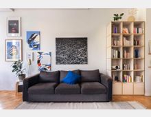 Wohnzimmer mit einer dunklen Sofa-Couch, dekoriert mit einem blauen Kissen. An der weißen Wand darüber hängen fünf gerahmte und ungerahmte Bilder in unterschiedlichen Größen, daneben ein Bücherregal mit offenen Fächern, gefüllt mit Büchern und Dekorationsgegenständen. Links steht eine Zimmerpflanze neben einer Stehleuchte.