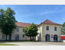 Ein zweistöckiges Gebäude mit hellbeiger Fassade und rotbraunem Ziegeldach, flankiert von zwei Bäumen. Die Fenster sind symmetrisch angeordnet, und der Eingangsbereich hat blaue Türrahmen mit Rundbögen; im Hintergrund ist eine gepflasterte Fläche zu sehen.