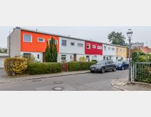 Reihenhaus-Siedlung mit hellen Fassaden in verschiedenen Farben (Orange, Weiß, Rot, Gelb), gepflegten Vorgärten mit Hecken und Bäumen, geparkten Autos an einer asphaltierten Straße und einem Straßenschild mit der Aufschrift „Lermooser Weg“ in einer Wohngegend.