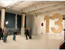 Fotostudio mit weißem Boden und Wänden, hohen Decken mit Lichtträgern und schwarzen Vorhängen an den Fenstern. Im Raum befinden sich große, goldene Ziffern „13“, mehrere Menschen, darunter ein Fotograf mit Kamera, sowie Beleuchtungsequipment.