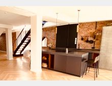 Moderne Wohnküche mit einer freistehenden Kücheninsel in dunklen Grautönen, einer Wand aus sichtbaren Ziegelsteinen und hängender Dekoration wie Töpfen und Pfannen. Im Hintergrund befindet sich eine schwarze Treppe mit Metallgeländer, die zu einem oberen Stockwerk führt, sowie ein helles Parkettbodenmuster.