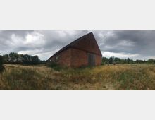 Eine alte Backsteinscheune mit schrägem Dach steht in einer überwucherten Wiesenlandschaft, umgeben von vereinzelten Bäumen und Gras. Der Himmel ist bewölkt, und im Hintergrund sind einige Personen sichtbar.