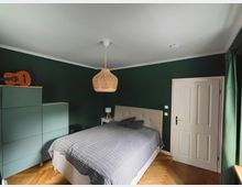 Schlafzimmer mit dunkelgrünen Wänden, Parkett im Fischgrätmuster und einem Doppelbett mit gepolstertem Kopfteil sowie grauer Tagesdecke. An der Decke hängt eine geflochtene Pendelleuchte; neben dem Bett stehen ein Nachttisch, links eine Kommode, rechts eine weiße Tür und ein Fenster mit grünen Vorhängen.