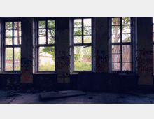 Verlassenes Gebäude mit großen, eingeteilten Fenstern, durch die eine überwucherte Außenlandschaft mit weiteren alten Ziegelgebäuden sichtbar ist. Die Wände im Inneren sind mit Graffiti bedeckt, der Boden ist teils beschädigt.