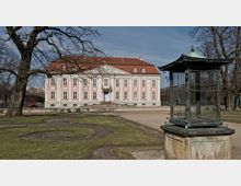 Barockes Gebäude mit rosa und weißen Fassaden, symmetrisch angeordneten Fenstern und einem roten Walmdach, inmitten eines Gartens mit Rasenflächen und Wegen. Im Vordergrund ein dekorativer, metallener Pavillon auf einem Steinsockel, umgeben von teils kahlen Bäumen.