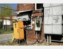 Eine Außenwand aus rotem Ziegelstein mit mehreren montierten Stromkästen, darunter ein gelber Verteilerkasten und eine transparente Abdeckung mit einem Stromzähler. Im Hintergrund sind ein Zaun und ein Schild mit der Aufschrift „Zu vermieten“ zu sehen.