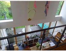 Wohnraum mit hohen Decken und großen Fensterfronten, die auf eine Terrasse mit Holzboden und Gartenblick führen. Im Innenraum sind ein Esstisch mit dekoriertem Geschirr, Pflanzen, Möbel sowie ein Klavier sichtbar.