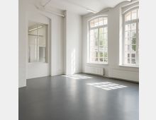 Heller, leerer Innenraum mit weißen Wänden, glattem grauen Boden und hohen, bogenförmigen Sprossenfenstern, durch die viel Tageslicht einfällt. An den Wänden befinden sich Heizkörper sowie ein verglastes Innenfenster zu einem Nebenraum.