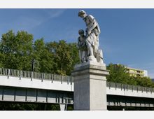 Steinfigurengruppe auf einem Sockel, die einen erwachsenen Mann und ein Kind darstellt, vor einer Baumreihe und einer Hochbahnbrücke aus Metall; im Hintergrund sind moderne Wohngebäude zu sehen.