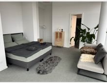 Ein modernes, helles Schlafzimmer mit einem grauen Polsterbett, grauer Bettwäsche und einem kleinen grauen Teppich auf einem Teppichboden. Im Raum befinden sich zudem ein kleines Holzschränkchen, ein Kleiderständer, eine graue Couch mit Kissen und eine große Pflanze neben einer weißen Wand. Eine Tür führt in ein weiteres Zimmer mit dunklen Fliesen.