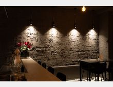 Dunkler Innenraum einer Bar oder eines Cafés mit einer strukturierten, beleuchteten Wand aus grau-verputztem Mauerwerk. Im Vordergrund ein Tresen mit Gläsern und einer Vase mit roten Blumen, daneben Tische mit schwarzen Stühlen.