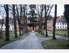 Pflasterweg, der von Bäumen gesäumt ist, führt zu einem großen, historischen Backsteingebäude mit roten Dachziegeln und weißen Fensterrahmen. Im Vordergrund befinden sich Rasenflächen, Gehwege und Parkschilder.
