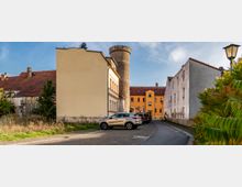 Eine enge Straße führt zu einem runden, historischen Wasserturm aus Stein, der von Wohnhäusern umgeben ist. Im Vordergrund sind geparkte Autos, eine Straßenlaterne und Büsche zu sehen, während die Fassaden der Gebäude teils renoviert und teils verwittert wirken.