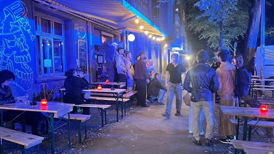 Außenbereich einer Bar an einer städtischen Straße bei Nacht, mit Holz-Bierzeltgarnituren und roten Kerzenlichtern unter einer Markise und Lichterkette. Die blau beleuchtete Fassade ist mit Graffiti versehen, daneben stehen Bäume und ein geparkter Transporter am Straßenrand.