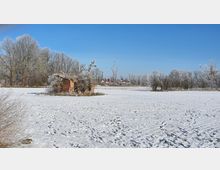 Schneebedecktes Feld mit einer kleinen, verfallenen Backsteinhütte im Vordergrund und kahlen, frostreichen Bäumen im Hintergrund unter klarem blauem Himmel. Im Hintergrund sind vereinzelt Häuser und weitere Bäume zu sehen.