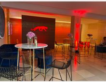 Ein moderner, eleganter Innenraum mit hohen Stehtischen und schwarzen Metallhockern, dekoriert mit Blumenarrangements in Glasvasen. Der Hintergrund ist in Rot- und Goldtönen beleuchtet, mit einer stilisierten Panther-Silhouette an der Wand.