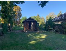 Ein kleiner Garten mit einer Holzlaube in der Mitte, umgeben von Rasenflächen, Bäumen und Sträuchern. Rechts steht ein einfacher Sitzbereich mit einer Bank und einem Tisch, während Zäune und Grünflächen den Garten umrahmen.