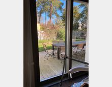 Blick durch eine Terrassentür auf eine Holzterrasse mit einem Holztisch und mehreren Metallstühlen. Im Hintergrund befindet sich ein Garten mit Rasen, Bäumen und Sträuchern.