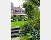 Zweistöckiges Wohnhaus mit roten Dachziegeln und weißen Fenstern, umgeben von einem gepflegten Garten mit grünem Rasen, Blumen, Sträuchern und Bäumen. Im Vordergrund führen Steinstufen durch eine mit Efeu und Farn bewachsene Böschung.