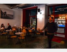 Innenraum einer modernen Bar mit einfacher Einrichtung, bestehend aus Holztischen und -stühlen. An der weißen Wand hängt ein großes Schwarz-Weiß-Foto, und durch die roten Fensterfronten ist eine beleuchtete Straße sichtbar; Kerzen auf den Tischen sorgen für eine gemütliche Beleuchtung.