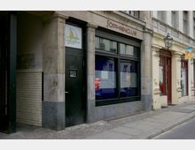 Außenansicht eines Gebäudes mit der Aufschrift „Sohienclub“ über einem schwarzen Eingang und dunkel getönten Fenstern, flankiert von einem weiteren Geschäftsportal mit heller Fassade und einer Laterne an der Wand. Der Gehweg davor ist aus Kopfsteinpflaster.