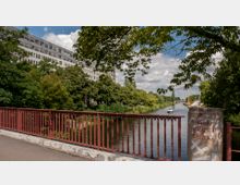 Ansicht von der Kaiser-Augusta-Brücke über einen von Bäumen gesäumten Kanal mit einem Boot. Im Hintergrund befindet sich ein modernes, graues Gebäude, während der Vordergrund von einem roten Metallgeländer und einer beschrifteten Brückenstütze dominiert wird.