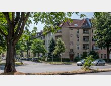 Straßenecke mit mehrstöckigem Wohnhaus im Hintergrund, das eine helle Fassade und rote Dachziegel hat. Umgeben von Bäumen, parkenden Autos und Straßenschildern in einem urbanen Wohngebiet.