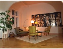 Wohnzimmer mit heller Holzbodenfläche, einer Sitzgruppe aus einem Sofa, zwei grünen Polstersesseln und einem kleinen Couchtisch auf einem Teppich. Im Hintergrund eine große Wandkunst mit Schwarz-Weiß-Motiven, eine Bogenlampe und ein großes Fenster; links steht eine Zimmerpflanze in einem Topf.