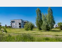 Moderner Wohnkomplex mit Flachdach und großen Fenstern, umgeben von einer weitläufigen, grünen Rasenfläche mit vereinzelten Bäumen und Sträuchern. Der Himmel ist klar und blau, typische ländliche Umgebung.