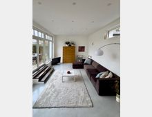 Helles, modern eingerichtetes Wohnzimmer mit hohen Decken, großflächiger Glasfront zur Terrasse und schmalen Oberlichtern an der rechten Wand. Im Raum stehen ein dunkles Ecksofa mit Bogenlampe, ein heller Teppich mit kleinem Couchtisch sowie ein hoher Holzschrank und ein niedriger Fernseher neben Stufen entlang der Fensterfront.