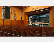 Innenraum eines Theatersaals mit roten Wänden und Holzakzenten. Im Vordergrund befinden sich Reihen von braunen Sitzplätzen, während im Hintergrund eine Bühne mit seitlichen Vorhängen, Beleuchtung und aufgebauten Kulissen zu sehen ist.