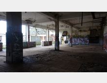 Innenraum eines verlassenen, heruntergekommenen Gebäudes mit Graffiti an den Wänden und Pfeilern. Der Raum zeigt freiliegende Rohre und Kabel an der Decke, beschädigte Fliesenböden und teils zerstörte Wände, mit Blick nach außen durch große Öffnungen.