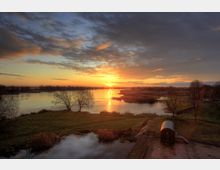 Sonnenuntergang über einem Fluss in einer ländlichen Landschaft mit vereinzelten Bäumen, offenem Wiesengelände und Wasserflächen. Im Vordergrund steht ein kleiner hölzerner Wagen auf Schienen, umgeben von Grasflächen und einem Picknicktisch.