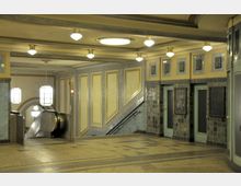 Unterirdischer U-Bahn-Bereich mit Jugendstil-Elementen, gekennzeichnet durch rechteckige Wandverkleidungen in Creme und Blau sowie eingebetteten Reliefs. Zu sehen sind eine Rolltreppe, Fliesenwände und moderne Pendelleuchten.