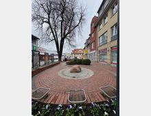 Eine kleine gepflasterte Platzfläche mit einem zentralen roten Stein auf einer runden, steinernen Einfassung, umgeben von roten Ziegeln. Im Hintergrund stehen ein großer, unbelaubter Baum, ein Einkaufszentrum und mehrstöckige Gebäude mit Geschäften.