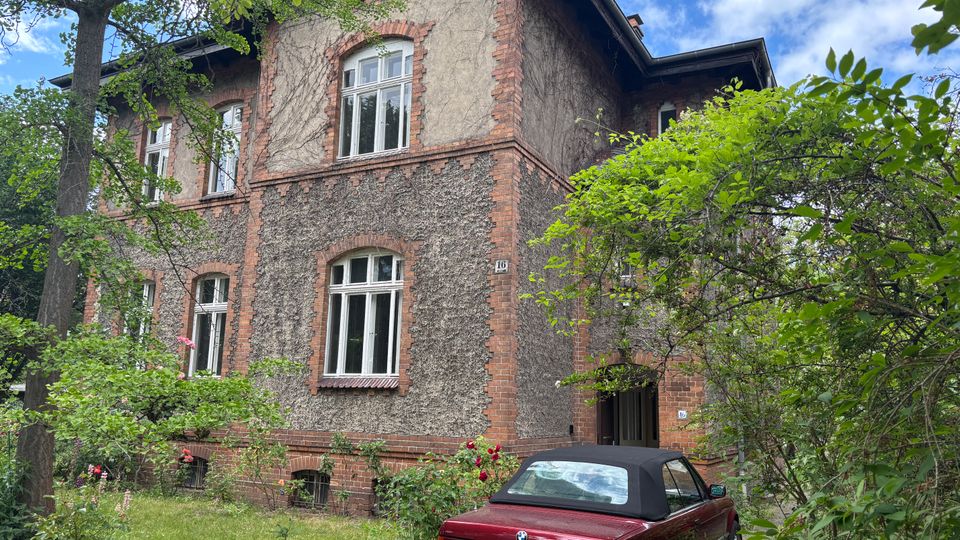 Mehrstöckiges Wohnhaus mit rau verputzter Fassade und roten Ziegelrahmen um die Fenster, teils mit Efeu bewachsen. Davor ein verwilderter Garten mit Bäumen und Sträuchern sowie ein geparktes rotes Cabrio nahe dem Hauseingang.
