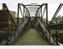 Eine historische Stahlbrücke mit Holzplanken und geschwungenen Ziergeländern führt über einen Fluss. Im Hintergrund sind Backsteingebäude mit dekorativen Fassaden und dahinter Bäume sichtbar.