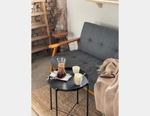 Gemütliche Sitzecke mit grauem Sofa, Holzlehnen und einer beigen Decke, daneben ein schwarzer runder Beistelltisch mit Karaffe, zwei weißen Tassen und einem Löffel. Im Hintergrund eine rustikale Backsteinwand, eine Holzleiter und ein kleiner Holztisch mit einer Vase und getrockneten Blumen.