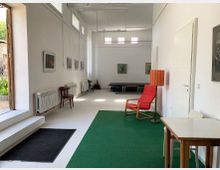 Ein heller, rechteckiger Raum mit weißen Wänden und hohen Fenstern. An der linken Wand hängen Bilder, im Vordergrund steht ein roter Sessel neben einer orangefarbenen Stehlampe auf grünem Teppich, während Heizkörper und Stühle entlang der Wände platziert sind.