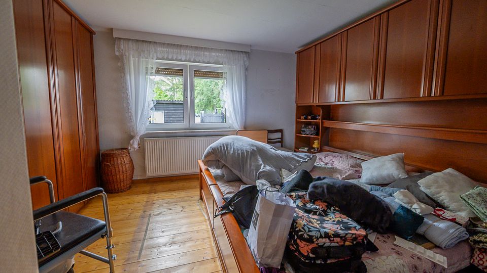 Schlafzimmer mit Holzfußboden, großem Bett und wandhohen Holzschränken an beiden Seiten. In der Mitte ein Fenster mit weißen Gardinen und Heizkörper darunter, daneben einfache Sitzmöbel und ein Korb.
