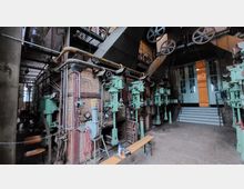 Industriehalle mit historischen Maschinen und Apparaturen, darunter grüne Metallanlagen und verlegte Rohre an einer Ziegelmauer. Im Hintergrund führt eine Treppe zu einer Tür mit gläsernen Fensterpaneelen.