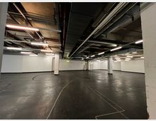 Ein leerer Innenraum mit schwarzem Boden, weißen Wänden und sichtbaren Rohrleitungen sowie Lüftungskanälen an der Decke. Der Raum ist beleuchtet durch rechteckige Leuchtstofflampen, und es sind mehrere Stützsäulen sowie Notausgangsschilder zu sehen.