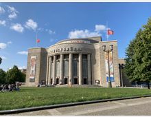 Außenansicht der Volksbühne Berlin, einem historischen Theatergebäude mit einer säulenförmigen Fassade aus hellem Stein. Im Vordergrund liegt eine Rasenfläche, die von Personen genutzt wird, und auf beiden Seiten des Gebäudes sind große Wandbilder angebracht.