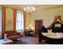 Historisches Schlafzimmer mit einem Himmelbett aus dunklem Holz, einer Sitzgruppe mit rotem Sofa, einem kleinen Schreibtisch und einem Kronleuchter. Die hohen Fenster sind mit Vorhängen versehen, und der Raum ist mit einem Orientteppich und klassischen Möbeln eingerichtet.