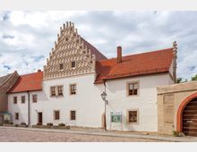 Historisches Gebäude mit weiß verputzter Fassade, roten Ziegeldächern und einem markanten, reich verzierten Stufengiebel. Vor dem Gebäude befinden sich Informationstafeln, Blumenkästen und eine gepflasterte Straße.