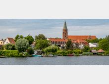 Stadtansicht mit einer Kirche mit Glockenturm und rotem Ziegeldach im Hintergrund, umgeben von Wohnhäusern und dichtem Grün, im Vordergrund ein Fluss mit Bootsanlegestellen und kleinen Booten.