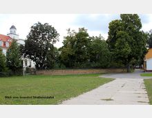 Blick auf einen Innenhof mit gepflastertem Weg, angrenzender Rasenfläche und Bäumen im Hintergrund. Links ist ein historisches Gebäude mit weißer Fassade und einem Turm erkennbar.
