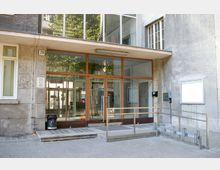 Eingangsbereich eines öffentlichen Gebäudes mit gläserner Doppeltür in einem modernen Rahmen aus Holz, umgeben von Betonwänden. Eine Rampe mit Geländer sowie ein Briefkasten und ein Aushang sind rechts vom Eingang angebracht.